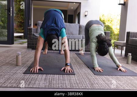 Ein Paar in Sportbekleidung übt Yoga im abwärts liegenden Hund auf überdachter Veranda mit schwarzen Matten Stockfoto