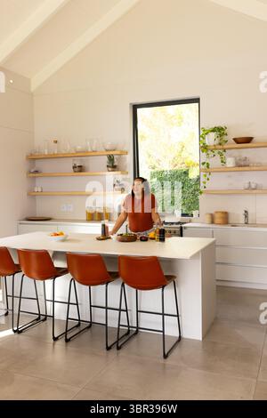 Asiatische Frau, die mit Händen auf der Kücheninsel steht, in der modernen Küche in der Nähe von Obstschalen Stockfoto