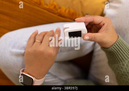 Asiatische Frau, die zu Hause auf dem Sofa sitzt und weißen Fingerspitzen-Pulsoximeter am linken Zeigefinger hält Stockfoto