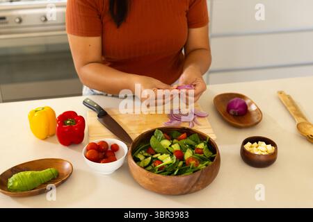 Asiatische Frau in der Küche schneidet rote Zwiebeln auf Schneidebrett und wirft Salat in Holzschüssel Stockfoto