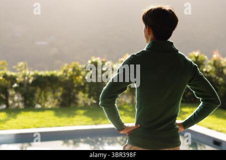 Asiatischer Mann, der mit Händen auf den Hüften am Pool steht, mit Blick auf Hecke, Rasen und Hügel, Kopierraum Stockfoto