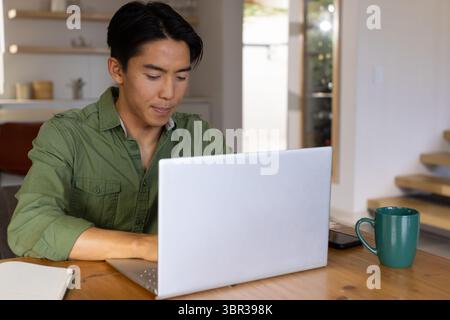 Asiatischer Mann, der am silbernen Laptop am Esstisch arbeitet, während er grünen Becher und Smartphone benutzt Stockfoto