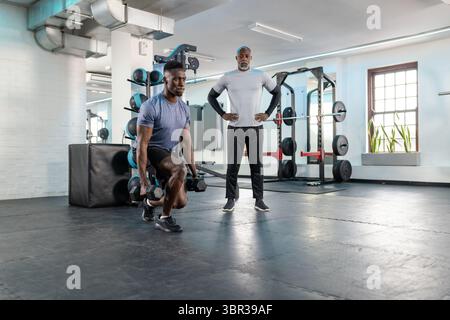 Afroamerikanischer Senior-Trainer, der einen erwachsenen männlichen Klienten trainiert, der einen Kurzhantel-Ausfallschritt im Fitnessstudio durchführt Stockfoto
