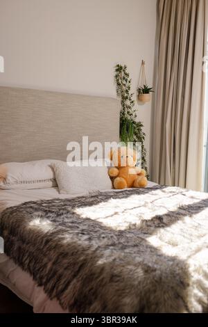 Queen-Size-Bett mit Kunstpelzüberwurf und Teddybären, Sonnenlicht strömt durch die Vorhänge Stockfoto
