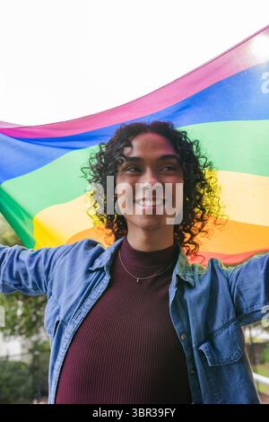 Nicht binäre Person mit großer Regenbogenfahne im Park lächelnd in Jeansjacke und kastanienbraunem Oberteil Stockfoto