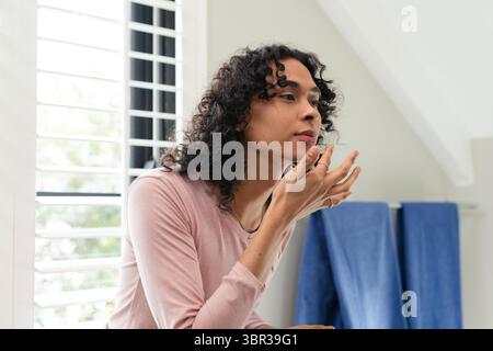 Nicht-binärer Erwachsener, der sich an den Schminktisch in der Nähe des verschlossenen Fensters lehnt und Serum neben blauen Handtüchern aufträgt Stockfoto