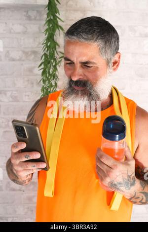 Lächelnder reifer Mann mit Tattoos, der ein orangenes Tanktop trägt und eine Wasserflasche hält, um nach dem Training sein Smartphone zu überprüfen Stockfoto