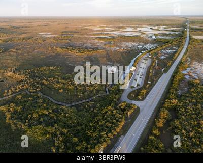 Panoramablick auf weite Feuchtgebiete, in denen sich windende Wasserstraßen das goldene Licht der Morgenröte reflektieren, im Kontrast zum dunkelgrünen Grün der dichten Wälder und einer geraden Straße, die durch die Landschaft führt, Everglades, Florida, USA Stockfoto