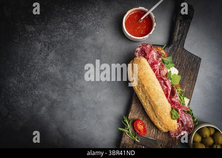 Salchichon extra oder Salami-Sandwich, gegrilltes Getreidebrot, mit Tomaten und Oliven auf dunklem Hintergrund, Draufsicht, Nahaufnahme. Gesundes hausgemachtes Frühstück Stockfoto