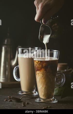 Frau Hand gießen Milch in Glas Tasse Kaffee auf schwarzem Hintergrund Stockfoto