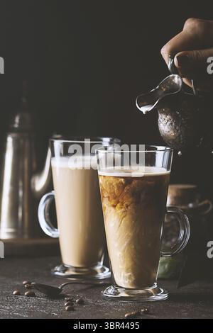 Frau Hand gießen Milch in Glas Tasse Kaffee auf schwarzem Hintergrund Stockfoto