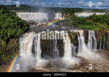 Blick auf majestätische Wasserfälle, die sich über felsige Klippen stürzen, inmitten von üppigem Grün, mit leuchtenden Regenbögen, die den Nebel in Iguazú, Ciudad Autónoma de BU, malen Stockfoto