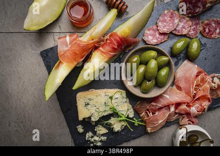 Schinken, Melonenscheiben, Brie- und Gorgonzola-Käse, Honig und Oliven auf grauem Hintergrund, Draufsicht Stockfoto