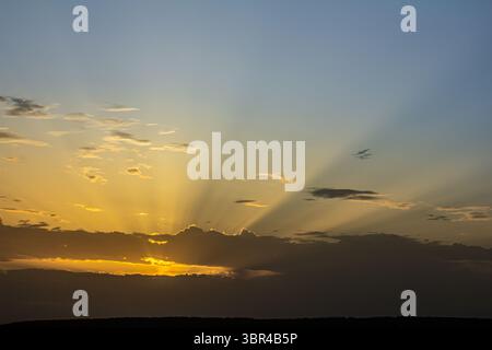 Der Himmel zeigt eine brillante Darstellung, während die Sonne untergeht und goldene Strahlen durch Wolken strömen und den Horizont in der Abenddämmerung erleuchten. Stockfoto