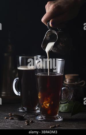 Frau Hand gießen Milch in Glas Tasse Kaffee auf schwarzem Hintergrund Stockfoto