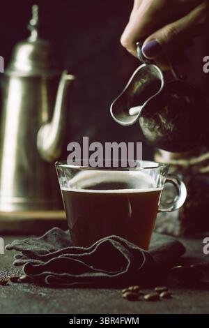 Frau Hand gießen Milch in Glas Tasse Kaffee auf schwarzem Hintergrund Stockfoto