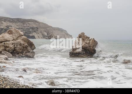 Blick auf raue, sonnenverwöhnte Felsen treffen auf das türkisfarbene Meer unter einem gedämpften Himmel, Wellen schlagen gegen die Küste in einer ruhigen Landschaft, Paphos, Paphos, Zypern. Stockfoto