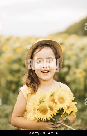 Ein lächelndes Mädchen mit Hut und gelbem Kleid mit Blumenstrauß auf dem Feld. Sommerurlaub Stockfoto