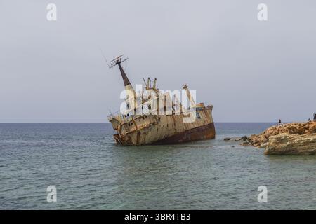 Paphos, Zypern - 25. Juni 2025: Blick auf das verrostete, verfallene Schiffswrack Edro III, das an der felsigen Küste ruht, ein krasser Kontrast zum ruhigen blauen Meer und dem bewölkten Himmel. Stockfoto