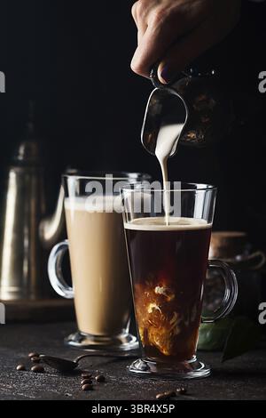 Frau Hand gießen Milch in Glas Tasse Kaffee auf schwarzem Hintergrund Stockfoto