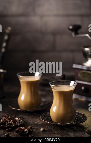 Tee oder Kaffee mit Milch und Gewürzen im Glas Armudu mit Kaffeebohnen und Zucker auf dunklem Hintergrund Stockfoto