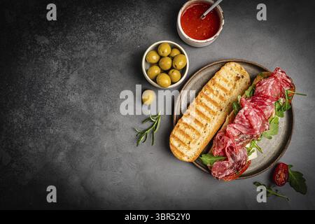 Salchichon extra oder Salami-Sandwich, gegrilltes Getreidebrot, mit Tomaten und Oliven auf dunklem Hintergrund, Draufsicht, Nahaufnahme. Gesundes hausgemachtes Frühstück Stockfoto