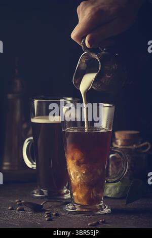 Frau Hand gießen Milch in Glas Tasse Kaffee auf schwarzem Hintergrund Stockfoto