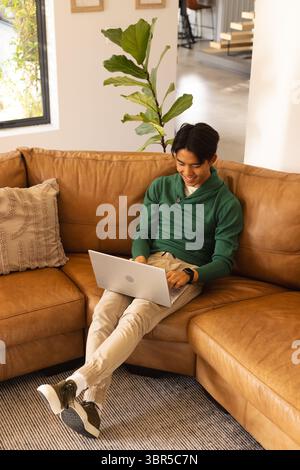 Asiatischer Mann, der auf dem Sofa sitzt und auf silbernem Laptop schreibt und zu Hause Smartwatch überprüft, Kopierraum Stockfoto