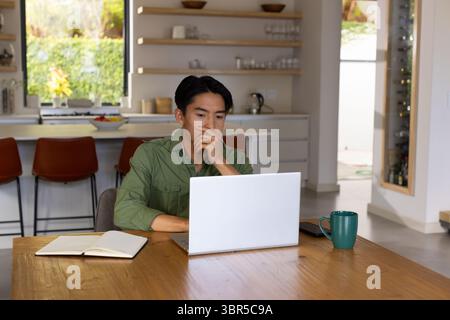 Asiatischer Mann, der am Esstisch in der Küche sitzt und auf silbernem Laptop mit dunkelgrünem Becher tippt Stockfoto