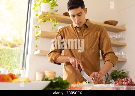 Asiatischer Mann, der Gemüse auf hölzernem Schneidebrett mit Kochmesser auf der Küchenzeile hackt Stockfoto