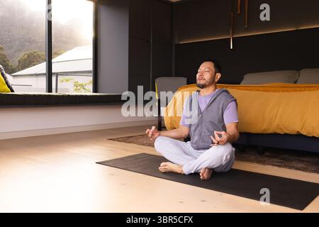 Erwachsener asiatischer Mann meditiert im Kreuz auf schwarzer Yogamatte in einem modernen Schlafzimmer am Fenster, Kopierraum Stockfoto