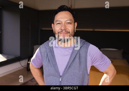 Asiatischer Mann, der im Schlafzimmer neben dem Plattformbett und der Bank steht, trägt einen grauen Hoodie und ein lila Hemd Stockfoto