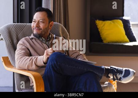 Asiatischer Mann, der eine Brille hält, während er im Holzsessel am Fenster sitzt, mit Notizbuch, Kopierraum Stockfoto