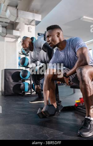 Zwei afroamerikanische Männer, die auf einer Bank im Fitnessstudio mit einem Medizinballständer gebogene Hantelreihen durchführen Stockfoto