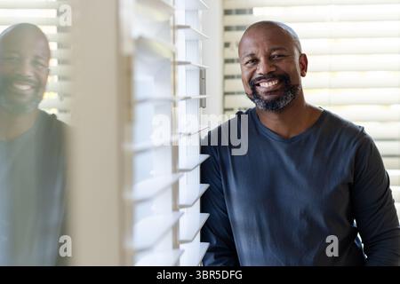 Reifer afroamerikanischer Mann, der sich zu Hause auf ein Fenster lehnt und sich in Glasscheibe hinter Jalousien reflektiert Stockfoto