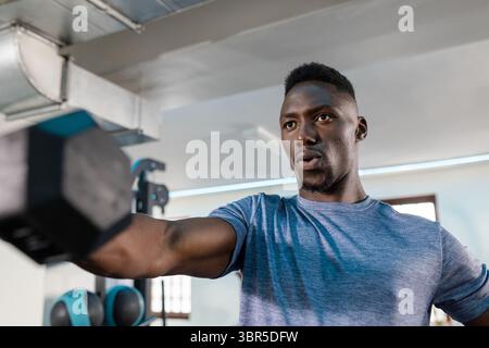 Ein afroamerikanischer Mann trägt ein blaues T-Shirt, der eine verstellbare Hantel im Fitnessstudio in der Nähe des Kettlebell-Gestells hebt Stockfoto