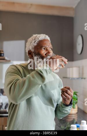 Ranghoher Afroamerikaner trinkt Wasser aus klarem Glas in moderner Küche mit Gläsern auf Regalen Stockfoto