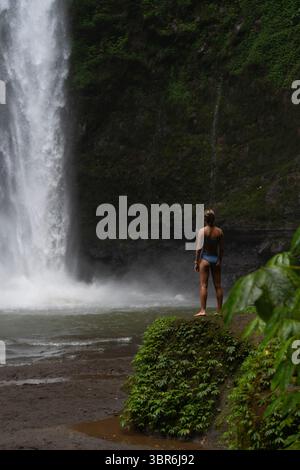Frau, die auf moosigem Felsen steht und auf den Dschungel-Wasserfall auf Bali blickt Stockfoto