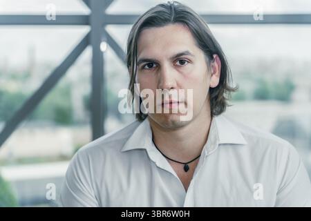 Porträt eines ernsten Mannes in einem weißen Hemd. Im Innenbereich, mit Fenster im Hintergrund. Stockfoto