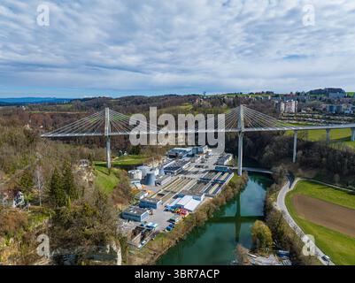 Luftaufnahme der Brücke Pont de la Poya, die sich anmutig über das Industriegebiet und den Fluss darunter erstreckt, unter einem Himmel verstreuter Wolken, Freiburg, Schweiz. Stockfoto