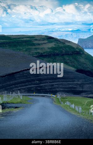 Blick auf die kurvenreiche Straße führt durch die raue, schwarze vulkanische Erde im Kontrast zu grünem Gras und fernen Klippen unter einem strukturierten Himmel, Vestmannaeyjabær, Vestmannaeyjabær, Island. Stockfoto