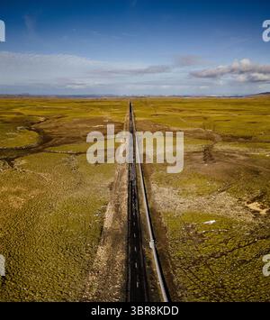 Aus der Vogelperspektive führt eine schroffe schwarze Straße durch die weitläufigen goldenen Felder unter einem riesigen Himmel, eine Studie in Kontrasten und abgelegener Schönheit, Mosfellsbær, Island. Stockfoto