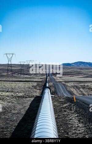 Blick auf eine silbrige Pipeline, die sich durch die trockene Landschaft erstreckt und parallel zu einer Straße unter einem klaren blauen Himmel verläuft, Nesjavellir, Grímsnes- og Grafningshreppur, Island. Stockfoto