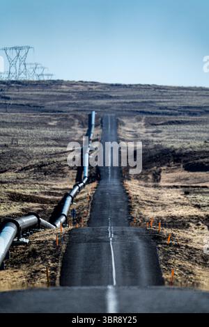Blick auf eine lange, schroffe Straße, die sich durch eine karge Landschaft erstreckt und mit industriellen Pipelines und entfernten Stromleitungen verbunden ist, Nesjavellir, Grímsnes- og Grafningshreppur, Island. Stockfoto