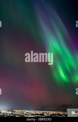 Blick auf die aurora Borealis tanzt in leuchtenden Grün- und Violetttönen über den funkelnden Lichtern der Stadt, einem himmlischen Ballett am Nachthimmel, Reykjavíkurborg, Reykjavík, Island. Stockfoto