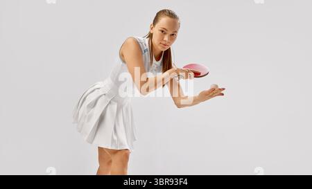Fokussierte Tischtennisspielerin, die Ball mit rotem Paddel auf neutralem Hintergrund serviert Stockfoto