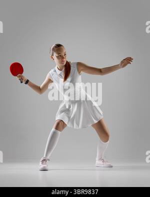Fokussierte Tischtennisspielerin in weißem Outfit bereitet sich auf grauen Hintergrund vor Stockfoto