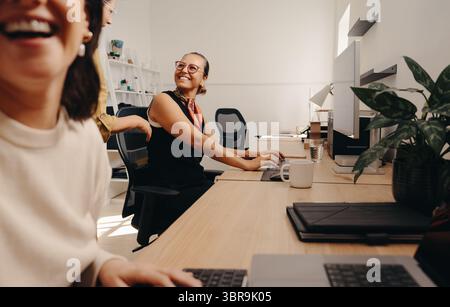 Helle Büroumgebung mit freundlichen Kollegen, die ein Lächeln teilen und in positiver Zusammenarbeit arbeiten. Optimistische und energiegeladene Atmosphäre Stockfoto