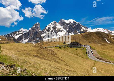 Die gewundene Straße des Passo di Giau führt zu einer Berghütte, auf der der schneebedeckte Gipfel des Antelao unter einem leuchtend blauen Himmel den Hintergrund dominiert. Stockfoto