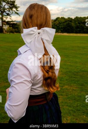 Von hinten sieht man eine Frau mit langen roten Haaren, die ein weißes Hemd mit einer großen weißen Schleife im Haar trägt. Sie steht auf einem grasbewachsenen Feld mit Tre Stockfoto